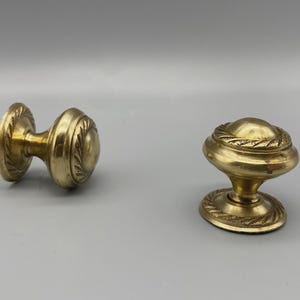 May include: Two antique brass door knobs. One is a double-sided knob with a decorative rope-like design around the edges. The other is a single knob with a similar design. Both are gold in color and have a vintage aesthetic.
