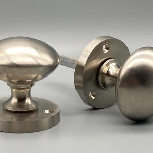 Op de afbeelding: Twee deurknoppen van geborsteld nikkel. Eén knop is losgemaakt en toont de montagehardware. De knoppen hebben een ovale vorm en zijn op een ronde basis gemonteerd. De deurbeslag is gemaakt van metaal en heeft een matte afwerking.