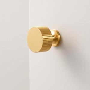 Op de afbeelding: Goudkleurige kastknop met een geribbelde, cilindrische handgreep en een cirkelvormige basis. De knop is bevestigd op een wit oppervlak, wat het minimalistische ontwerp benadrukt. Geschikt voor laden en kasten.