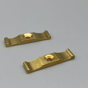 May include: Two small, brass-coloured metal brackets with a wavy design. Each bracket has a central hole and curved ends. The brackets are displayed on a light grey background.