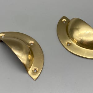 Può includere: Due maniglie a conchiglia in ottone lucido, ciascuna con un design a forma di semicerchio e tre fori per le viti. L'hardware ha una finitura brillante e riflettente, che suggerisce un'estetica vintage o antica. Ideale per aggiornamenti di armadi o cassetti.