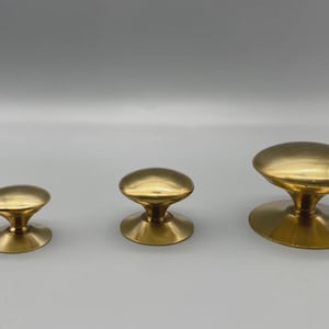 May include: Three brass cabinet knobs in varying sizes. Each knob features a rounded, oval-shaped top and a flared base. The knobs are arranged from smallest to largest, showcasing their different dimensions and design.