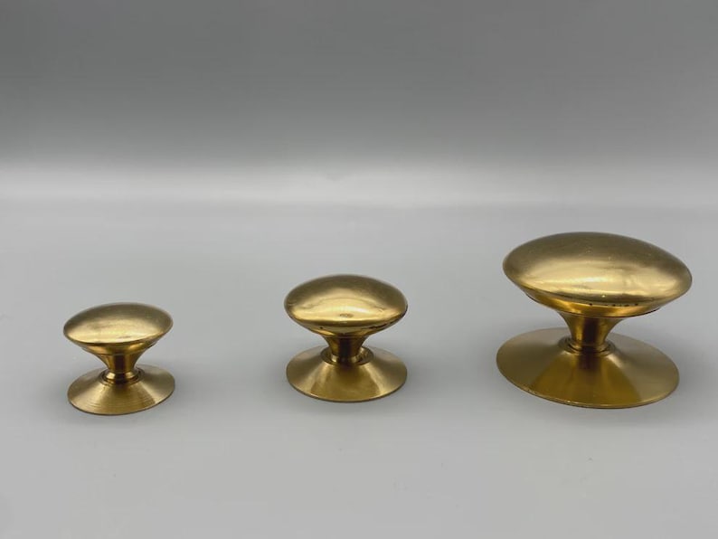 May include: Three brass cabinet knobs in varying sizes. Each knob features a rounded, oval-shaped top and a flared base. The knobs are arranged from smallest to largest, showcasing their different dimensions and design.