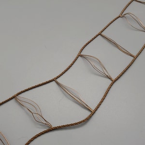May include: A decorative, openwork design featuring a series of interconnected, leaf-shaped elements. The design is constructed from a light brown, braided cord, with delicate, translucent, leaf-like accents. The overall aesthetic is delicate and airy.