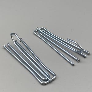 May include: Two silver curtain hooks. Each hook features a series of parallel rods with rounded ends, designed for hanging curtains. The hooks are made of metal and are set against a neutral grey background.