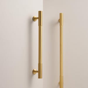 May include: Two gold door handles fixed to a white wall. The handles are long, cylindrical, and have a ribbed texture at the top and bottom. The handles are attached to the wall with round, gold-coloured mounts.