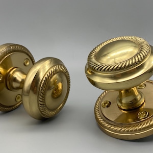 Op de afbeelding: Twee antieke messing deurknoppen. De knoppen hebben een ronde vorm met een decoratieve touwachtige rand. De basisplaten zijn ook rond met een bijpassend touwontwerp. De hardware heeft een glanzende goudkleur.