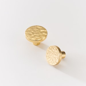 May include: Two gold-colored cabinet knobs with a hammered texture. The knobs have a circular design with a short, cylindrical stem. The knobs are made of metal and are set against a white background. These knobs are suitable for furniture and home decor.