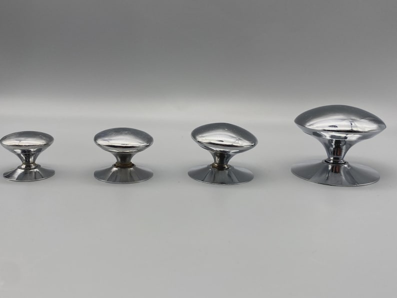 May include: Four shiny, chrome-finished cabinet knobs of varying sizes are arranged in a row. Each knob features a rounded, oval-shaped top and a flared base. The knobs are designed for furniture or cabinetry.