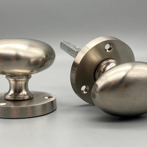 Può includere: Due pomelli per porte in nichel spazzolato. Ogni pomello ha una forma ovale ed è montato su una base rotonda. Un pomello è smontato, mostrando il mandrino. L'hardware è progettato per porte interne.