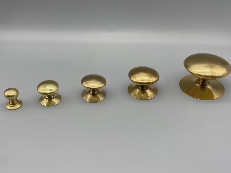May include: Five brass cabinet knobs of varying sizes are arranged in a row. The knobs have a rounded top and a circular base. The knobs are a shiny gold color and are set against a neutral background.