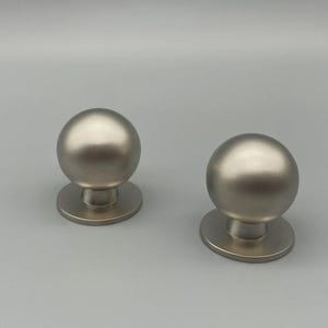 May include: Two brushed nickel door knobs with a spherical design. Each knob has a round base and a smooth, metallic finish. The knobs are displayed against a light gray background, showcasing their simple, modern aesthetic.
