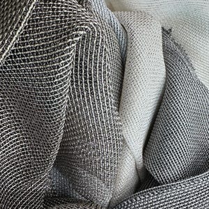 May include: Close-up of folded fabric in shades of grey and white. The fabric has a textured, woven appearance with a grid-like pattern. The folds create shadows and depth, highlighting the different tones and textures of the material.