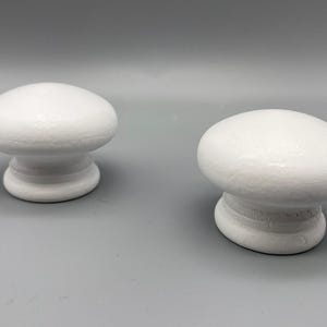 May include: Two white, oval cabinet knobs with a textured finish. Each knob has a rounded top and a cylindrical base. The knobs are set against a grey background, showcasing their simple design. These knobs are ideal for adding a touch of minimalist style to any furniture.