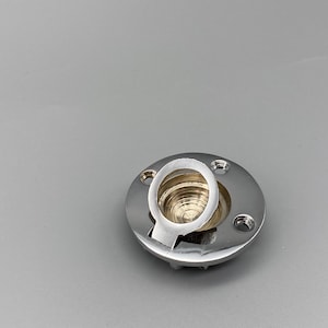 Puede incluir: Una pieza metálica plateada y brillante, con una base circular y un elemento en forma de anillo elevado. La base tiene varios agujeros pequeños alrededor de su perímetro. El anillo central tiene una ranura y un interior texturizado.