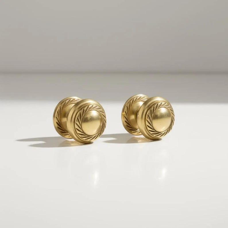 May include: Four brass cabinet knobs. Each knob features a circular design with a central disc and a detailed, textured border. The knobs are a warm gold colour and are arranged in two pairs.