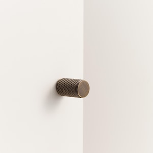 May include: A bronze cabinet knob, featuring a textured cylindrical body and a flat, circular face. The knob is mounted on a white surface. This hardware piece is designed to enhance the aesthetic of any cabinet or drawer, adding a touch of sophistication.