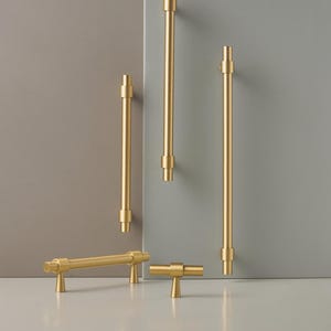 May include: A selection of gold-toned cabinet handles and knobs. The image shows various sizes of bar pulls and T-shaped knobs. The hardware has a modern, minimalist design. The items are displayed against a neutral grey and beige backdrop, emphasizing the metallic finish.