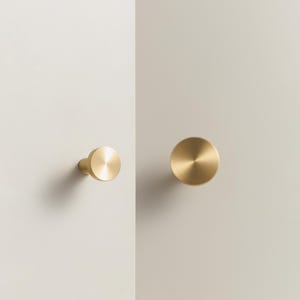 May include: Two brass cabinet knobs. One knob is attached to a white surface, while the other is detached. The knobs are circular with a brushed finish. The knobs are a warm gold color and are set against a neutral background.