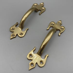 Könnte beinhalten: Zwei goldfarbene Messing-Schrankgriffe mit geschwungenem Griff und dekorativen Enden. Jeder Griff hat drei Befestigungslöcher und ein einzigartiges, verziertes Design.