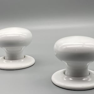 May include: Two white ceramic door knobs with a rounded top and a circular base. The knobs are glossy and appear to be new. The background is a solid light gray.