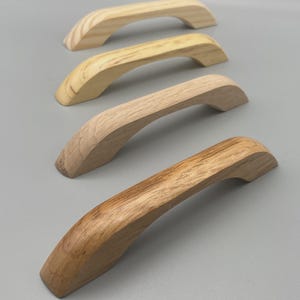 May include: Four wooden cabinet handles in different shades of natural wood. The handles are curved with a smooth finish, showing the wood grain. These handles are designed for furniture and home decor projects.