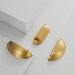 May include: Three gold-coloured, half-moon shaped cabinet pulls are arranged on a white surface. The pulls have a smooth, curved design and a shiny finish. The hardware is likely for drawers or cabinets.