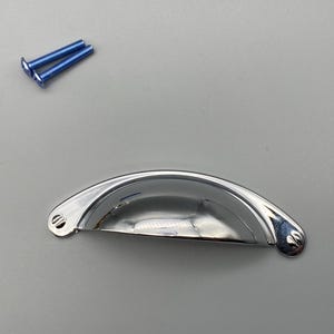Peut inclure: Une poignée de meuble en forme de coupe, chromée et brillante, avec deux vis bleues. La poignée est incurvée et a une surface réfléchissante. Idéale pour les armoires de cuisine ou de salle de bain.