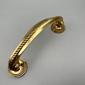 Può includere: Una maniglia dorata per mobili con manico curvo e dettagli decorativi a corda. La maniglia ha due piastre di montaggio circolari con fori per le viti. L'hardware è probabilmente in ottone o metallo simile.