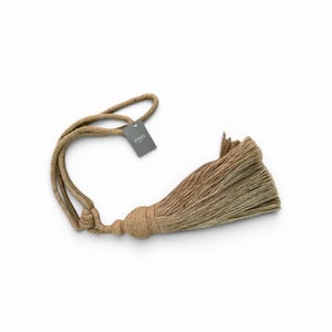 Op de afbeelding: Een beige kwast met een lusvormig touw en een label met de tekst "JONES". De kwast is gemaakt van meerdere strengen beige materiaal, met een afgeronde basis en een lang, vloeiend uiteinde. Het touw is bij de kwast geknoopt.