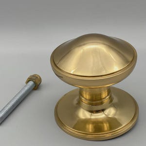 May include: A gold-coloured door knob with a rounded top and a circular base. A silver bolt with a nut is next to the knob. The knob has a shiny, reflective surface. Door hardware.