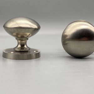 May include: Two brushed nickel door knobs. One is oval-shaped, mounted on a circular base with screw holes. The other is a spherical knob. Both have a metallic sheen against a neutral background.