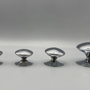 May include: Four chrome cabinet knobs of varying sizes are arranged in a row. Each knob has a rounded, oval-shaped top and a circular base. The knobs are reflective, with a shiny, metallic finish. They are likely for furniture or cabinetry.