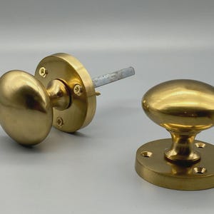 May include: Two brass door knobs. One knob is attached to a circular plate with screws and a metal rod. The other knob is detached and sits on a circular base with screw holes. Both knobs have an oval shape.