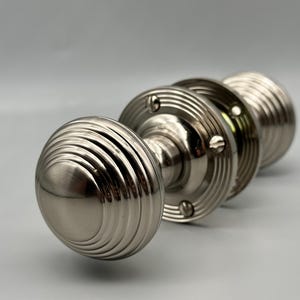 Könnte beinhalten: Ein Satz Türknöpfe aus gebürstetem Nickel. Jeder Knopf hat ein abgerundetes, gerilltes Design. Die Knöpfe sind an einer kreisförmigen Basis mit Schraubendetails befestigt. Die Hardware wird vor einem hellgrauen Hintergrund präsentiert.