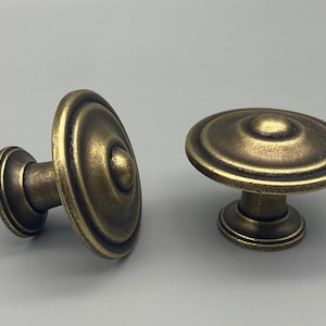 May include: Two antique-style cabinet knobs with a bronze finish. The knobs have a circular, layered design with a central raised button. One knob is on its side, and the other is upright.