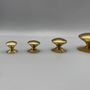 May include: Five brass cabinet knobs in varying sizes are arranged in a row. The knobs have a mushroom-shaped design with a rounded top and a flared base. The finish is a shiny, polished gold.