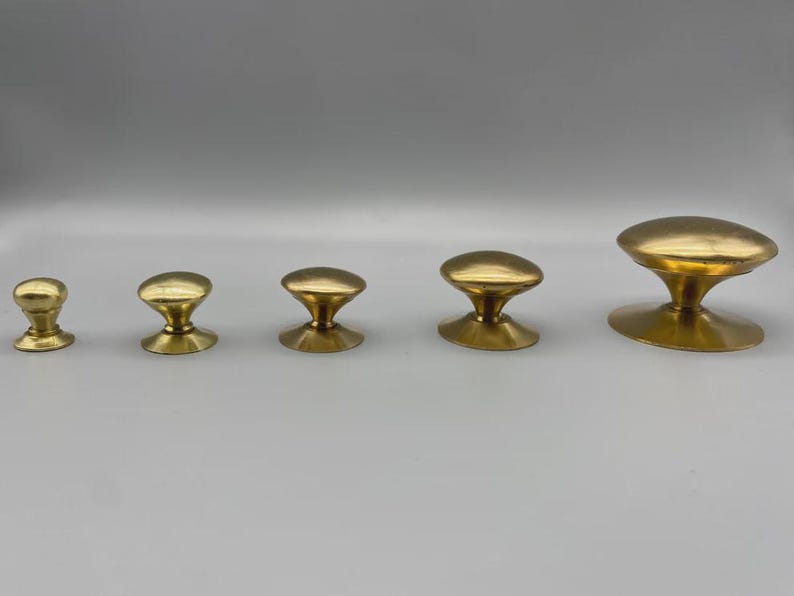 May include: Five brass cabinet knobs in varying sizes are arranged in a row. The knobs have a mushroom-shaped design with a rounded top and a flared base. The finish is a shiny, polished gold.