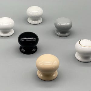 May include: A collection of six ceramic cabinet knobs in various colors, including white, black, gray, and beige. One white knob has a gold accent. The knobs are round with a pedestal base.