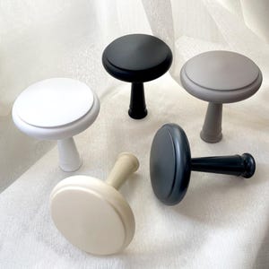 Może przedstawiać: Pięć dekoracyjnych gałek do szafek w różnych kolorach: białym, czarnym, szarym i beżowym. Każda gałka ma okrągłą górę i cylindryczny trzon. Gałki są ułożone na białej, teksturowanej powierzchni.