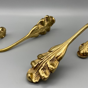 May include: Two ornate, gold-coloured curtain tiebacks. Each tieback features a curved hook design with a decorative leaf-like embellishment. The tiebacks are made of a shiny, metallic material and are set against a neutral background.