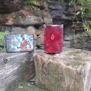 May include: Two items rest on a weathered wooden surface. One is a rectangular card with an illustration of a figure holding a weapon, with text that reads "OF LIGHTNING". The other is a rectangular, translucent, red object.
