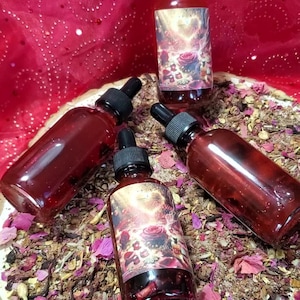 May include: Four glass dropper bottles filled with a deep red liquid, arranged on a bed of dried herbs and flower petals. The bottles have black caps and labels with a floral design. The background is a red fabric.