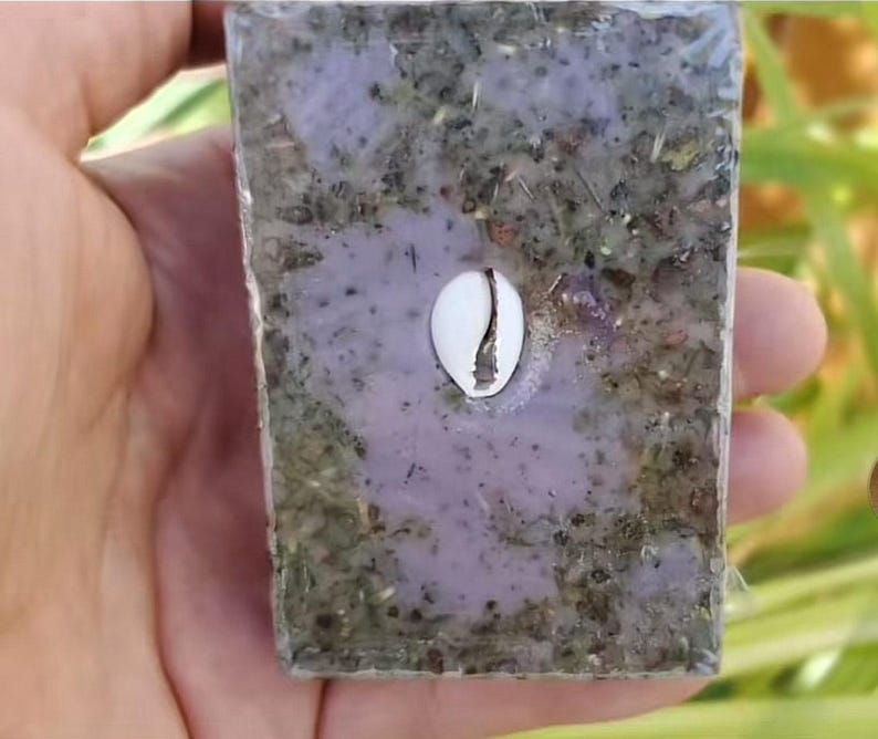 Puede incluir: Una pastilla de jab&oacute;n rectangular con un centro morado claro y una capa exterior moteada verde y marr&oacute;n. Una concha de cauri blanca est&aacute; incrustada en el centro. El jab&oacute;n se sostiene en una mano.