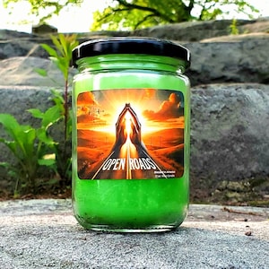 May include: A green candle in a glass jar with a black lid. The label shows hands reaching towards a road with the words "OPEN ROADS". The candle is 340 grams.
