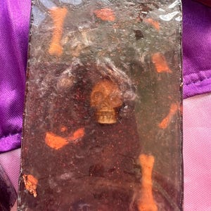 Peut inclure: Un pain de savon rectangulaire avec une base marron foncé, incrusté d'un petit crâne et d'os. Le savon a une couche supérieure claire et légèrement mousseuse, et des paillettes orange.