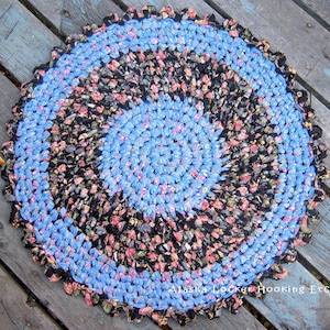 Könnte beinhalten: Ein runder, gehäkelter Teppich mit einem blauen und schwarzen Blumenmuster. Der Teppich ist aus recycelten Stoffresten gefertigt und hat einen gewellten Rand.