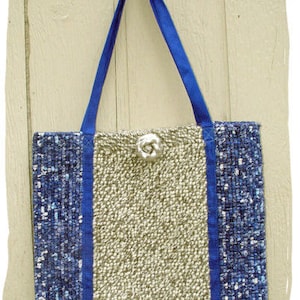 May include: A woven tote bag with blue and white patterned fabric on the sides and a beige woven fabric in the center. The bag has blue straps and a silver flower embellishment.