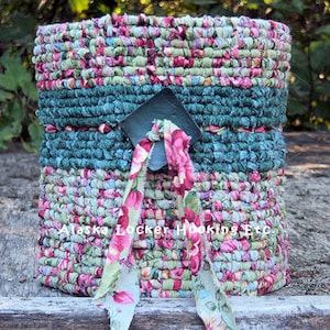May include: A round woven basket with a floral pattern in shades of pink, green, and blue. The basket has a green fabric band around the middle and a metal tag with the text "Alaska Locker Hooking Etc."