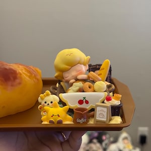 May include: A miniature figurine set on a brown tray, featuring a sleeping figure with yellow hair surrounded by miniature baked goods, coffee cups, and cartoon characters. A large, yellow pastry is on the left.